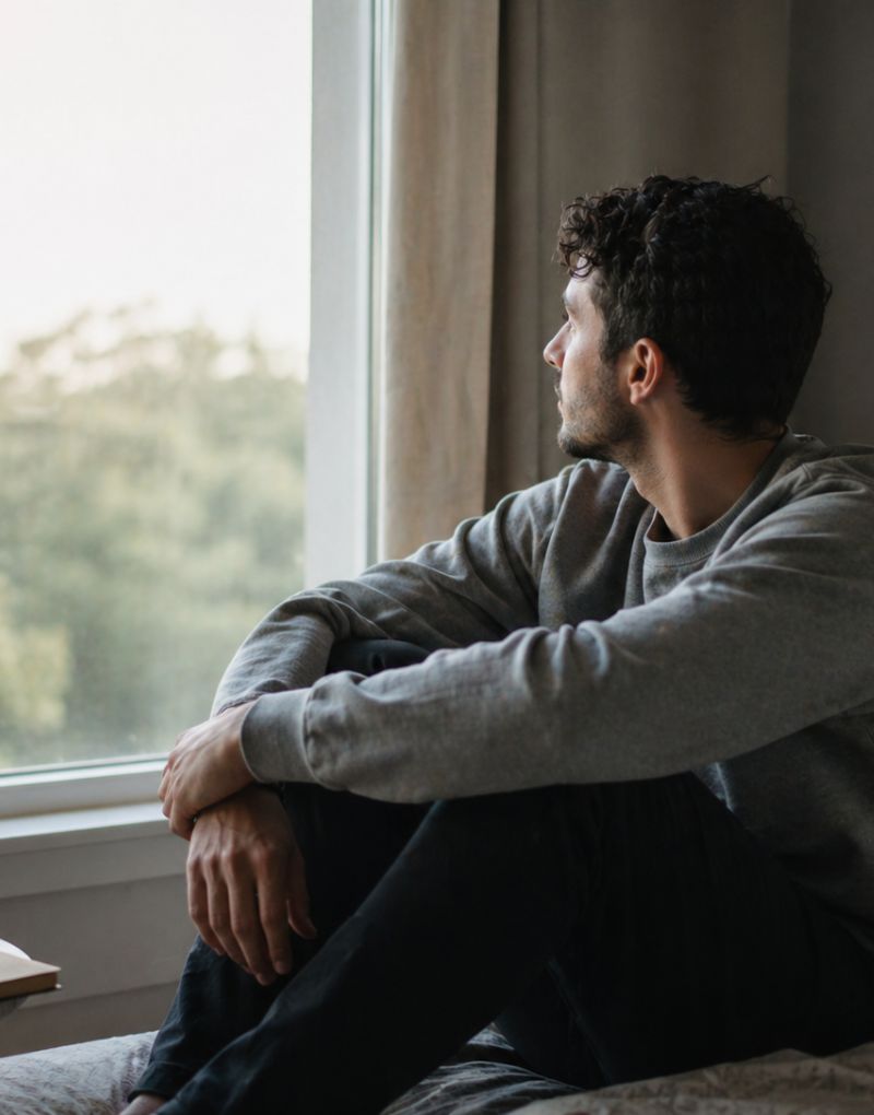 Hombre sentado junto a una ventana mirando hacia afuera en un momento de introspección y calma, representando el acompañamiento emocional